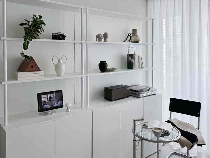 Minimal white-toned living room featuring clean bookshelf and transparent side table with coffee and book for a cozy atmosphere