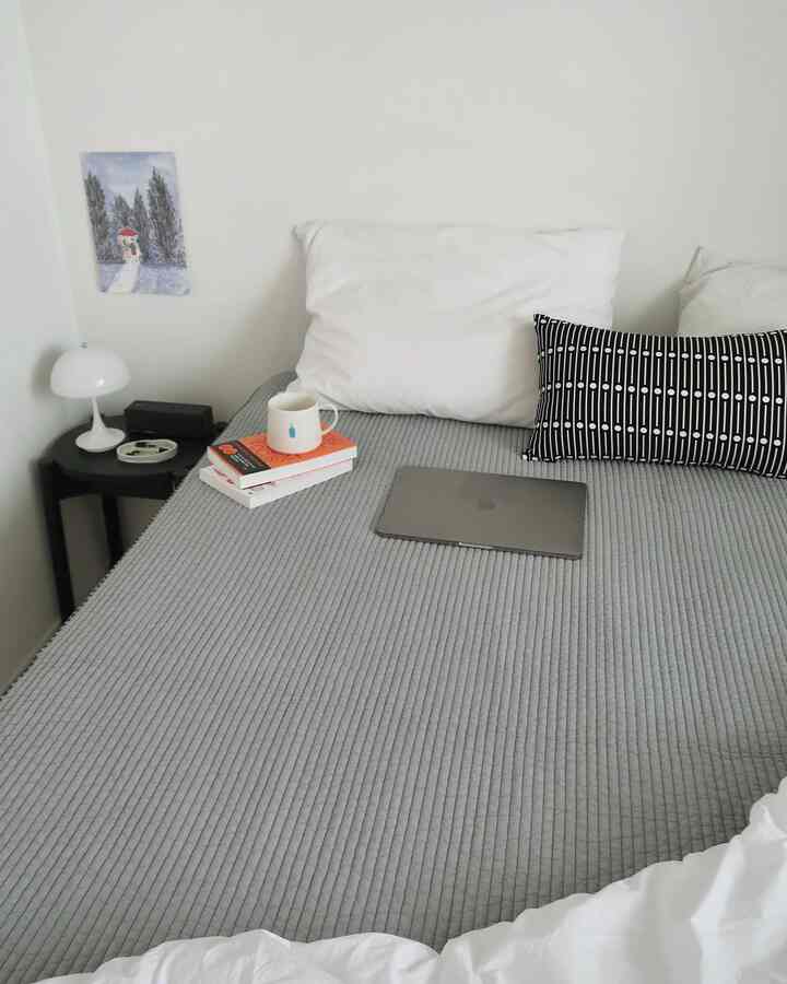 グレーと白のトーンの寝室、キルティングのベッドカバーとクッション、テーブルランプとノートパソコンが配置された清潔感のある空間