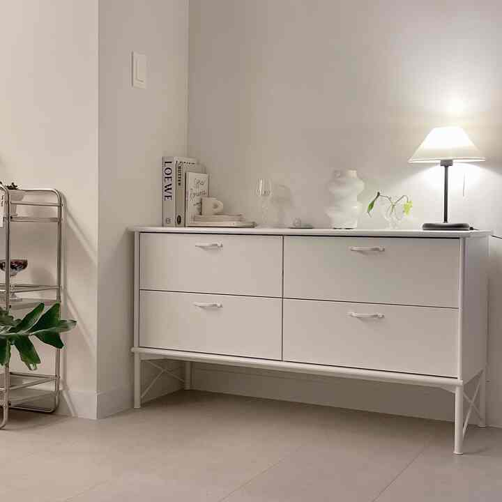 柔らかな白いトーンのリビング、白い4段のチェスト、テーブルランプ、ヴィンテージのガラスキャンドルホルダーが配置された洗練された空間