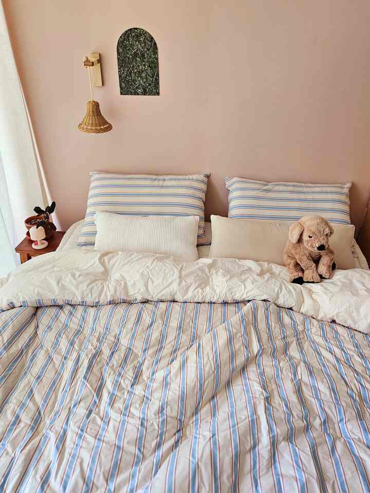 淡いピンク色の壁とブラウンのランプ、ストライプの寝具、ぬいぐるみの犬が置かれた温かみのある寝室