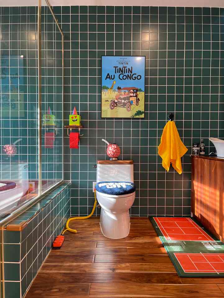 Vintage-style bathroom with green tiled walls and wood-tone floor, featuring a centered toilet and colorful accent accessories