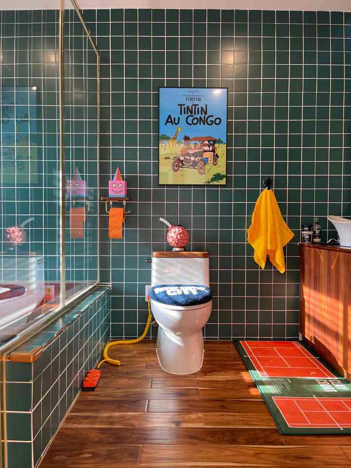 Retro-style bathroom with deep green tiled walls and warm wood flooring, featuring a white toilet centered below a vintage framed poster