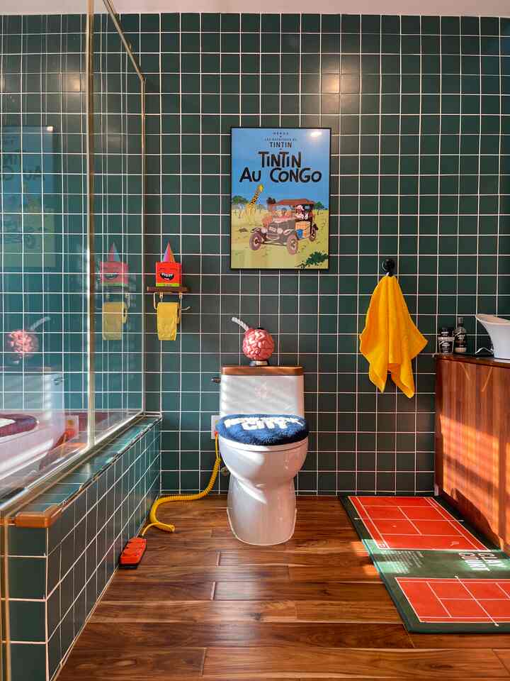 Green tiled wall and wood-toned floor bathroom featuring a toilet centered below a Tintin poster, accented with a yellow towel and bath mat