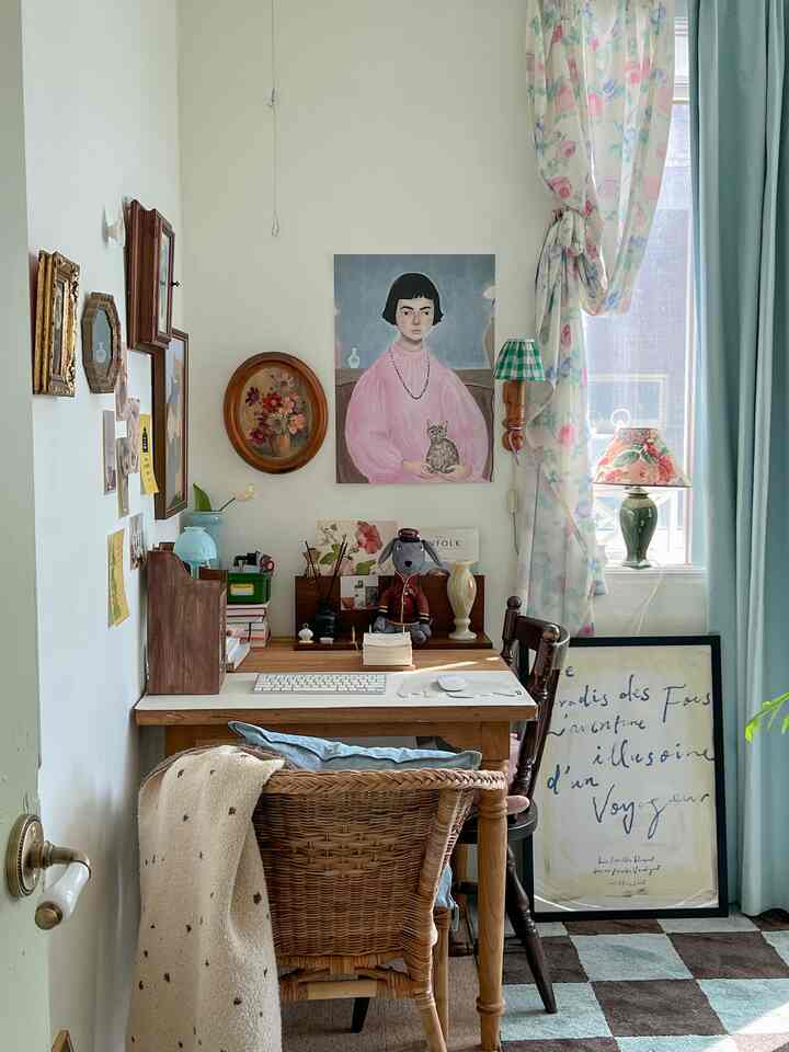 Natural wood-toned small home office featuring a wooden desk and wicker chair, framed artworks on wall, floral curtains creating cozy atmosphere
