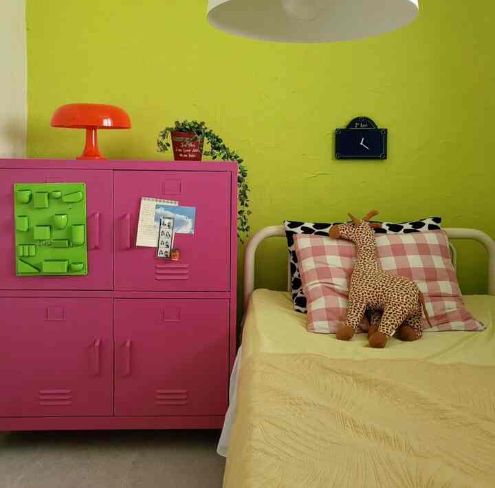 ピンクのキャビネットと黄色の壁が調和する子供部屋、ベッドにはキリンのぬいぐるみとチェック柄のクッションが置かれた明るいインテリア