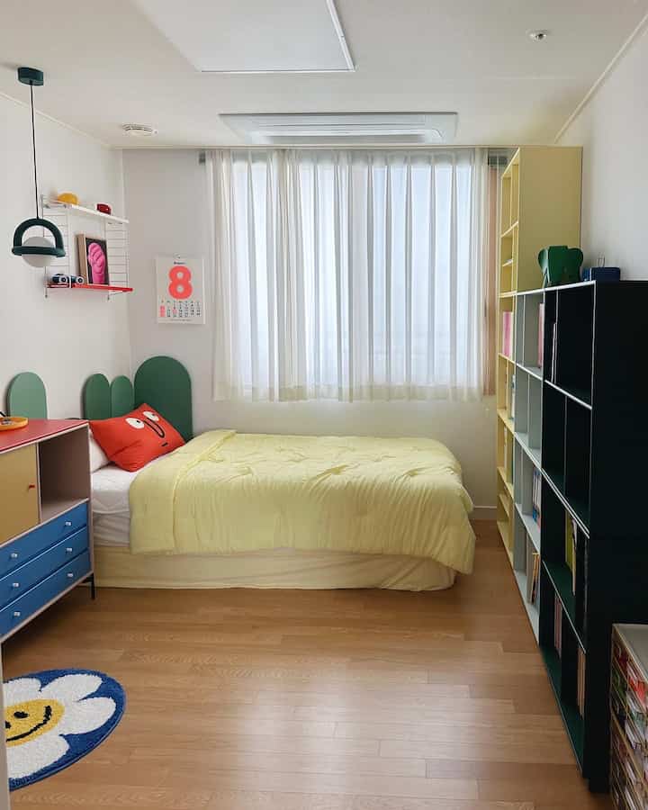 Bright kids' room with natural wood flooring and white walls, featuring light yellow bedding and colorful bookshelves creating a cozy atmosphere