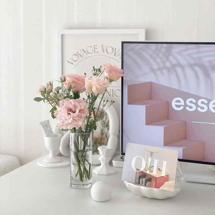 ホワイトトーンのデスクスペース、ピンクの花束とモニター、詩的なポスターが配置されたシンプルなホームオフィスインテリア