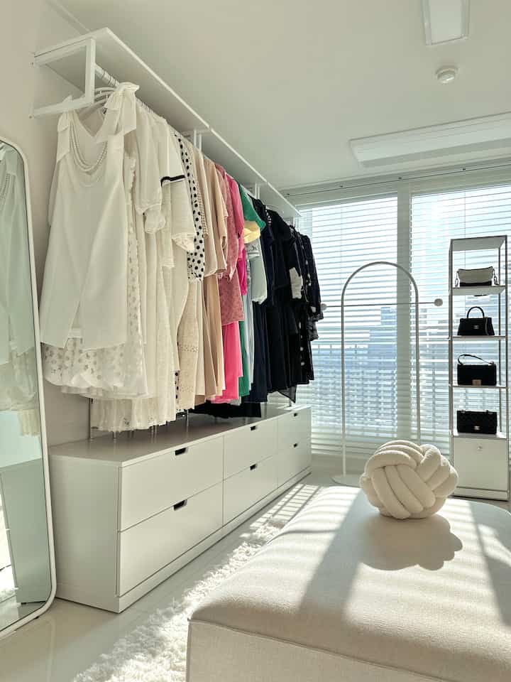White-toned walk-in closet space with a long wardrobe on left, beige bench, and window blinds creating a bright atmosphere