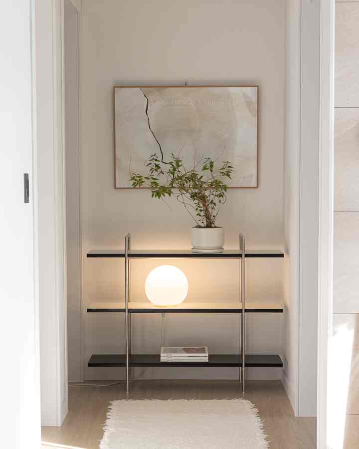 ナチュラルトーンの狭い玄関空間、中央に黒い棚があり、植物と丸い照明が配置されたシンプルなインテリア