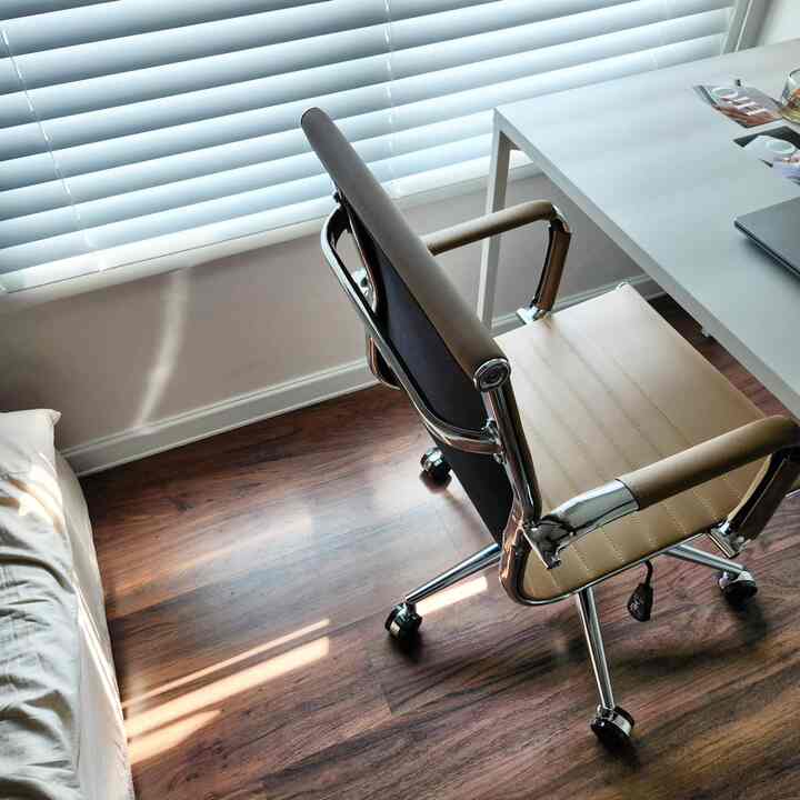 Small home office with white blinds and natural wood tone flooring, featuring beige office chair and white desk