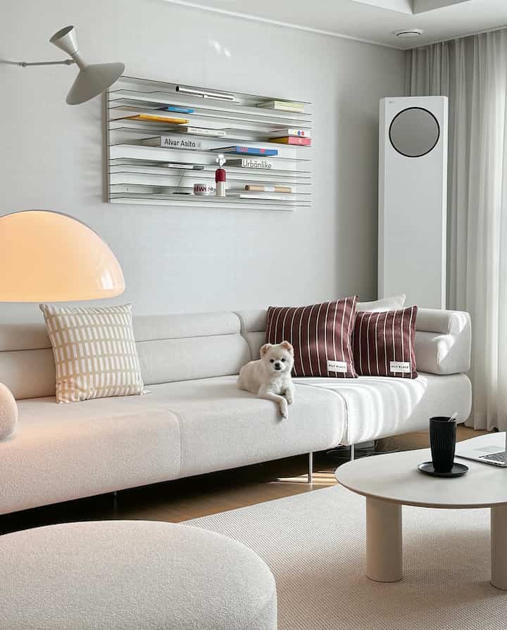 White and beige toned living room featuring large sofa, coffee table, floor lamp, and a small dog creating cozy atmosphere