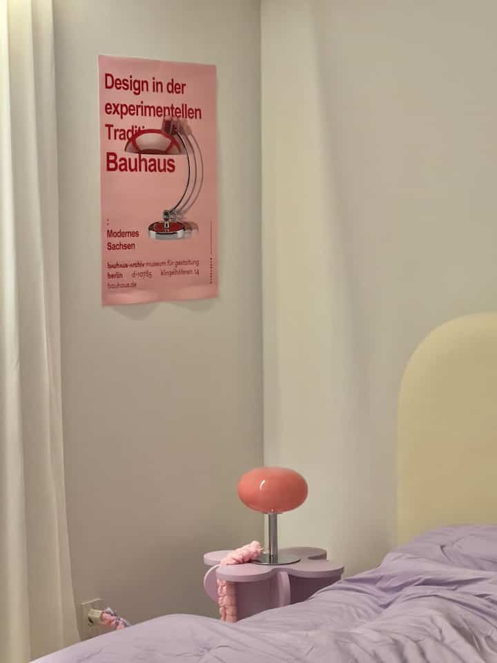 柔らかなピンクと紫の色調の寝室インテリア、花の形のナイトテーブルとミッドセンチュリーモダン風のポスターが配置された落ち着いた空間