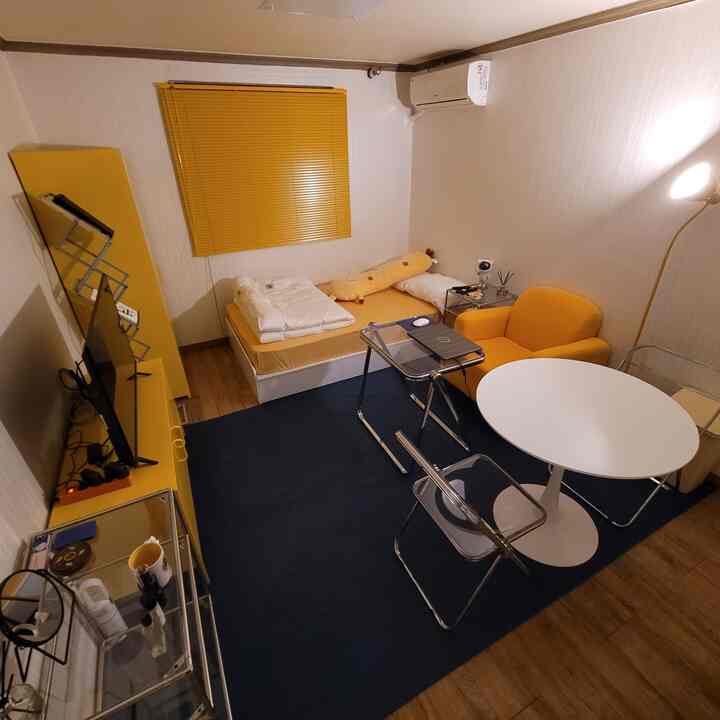 明るい黄色調のワンルーム、黄色のソファとブラインド、白い丸テーブルと透明な椅子が配置されたモダンなインテリア