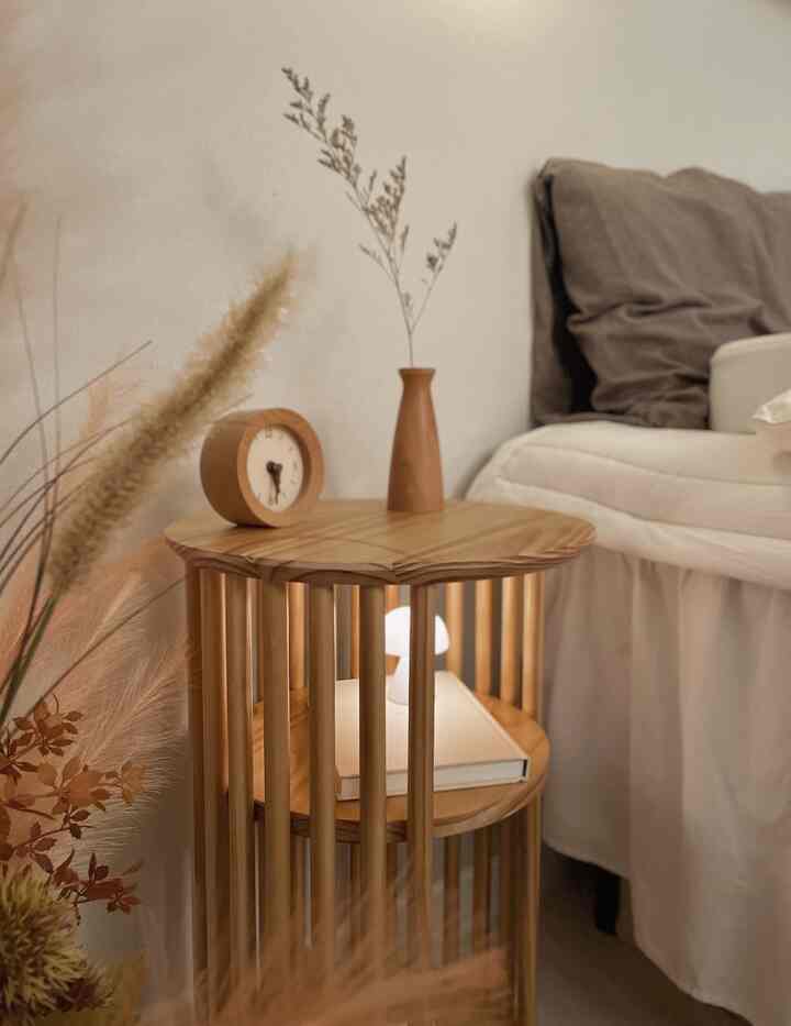 ナチュラルな木目調と白い寝具の調和が魅力的な寝室の隅、木製の円形サイドテーブルに花瓶とアナログ時計が配置された温かみのある空間