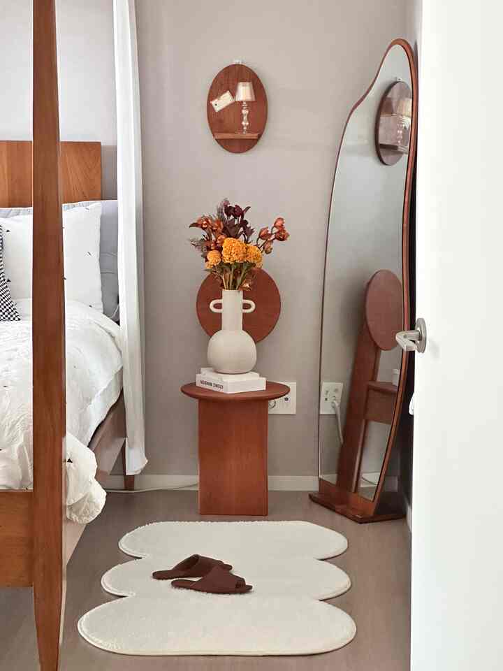 Cozy bedroom featuring wood tones and white with a nightstand holding a vase of flowers and a curved full-length mirror in a natural style space