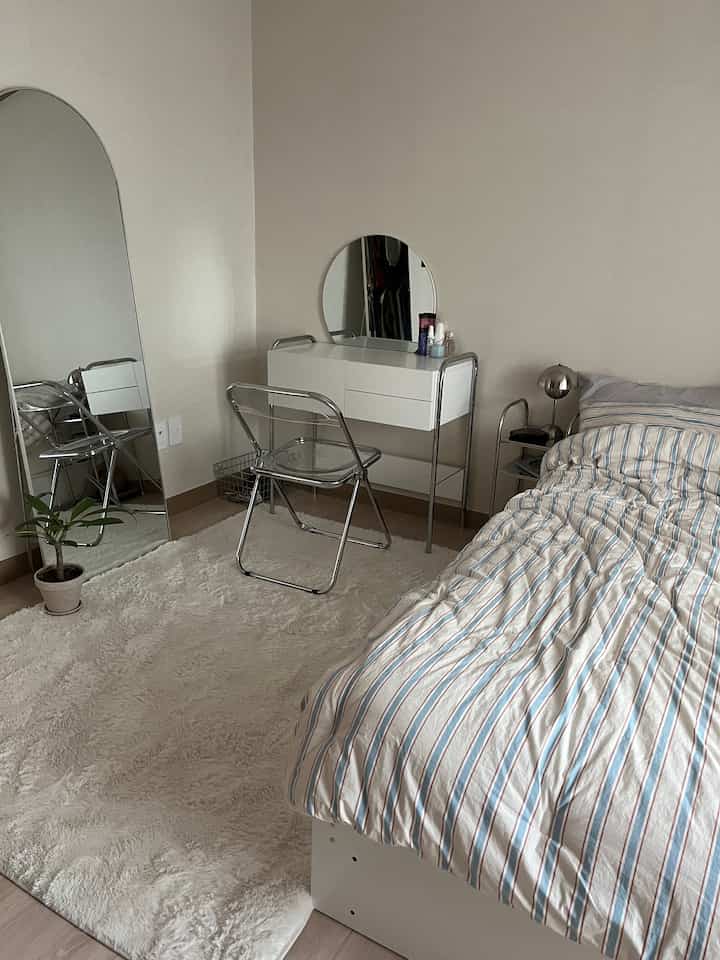 White-toned small and compact bedroom featuring a transparent chair, vanity with round mirror, and large rug in a simple setting