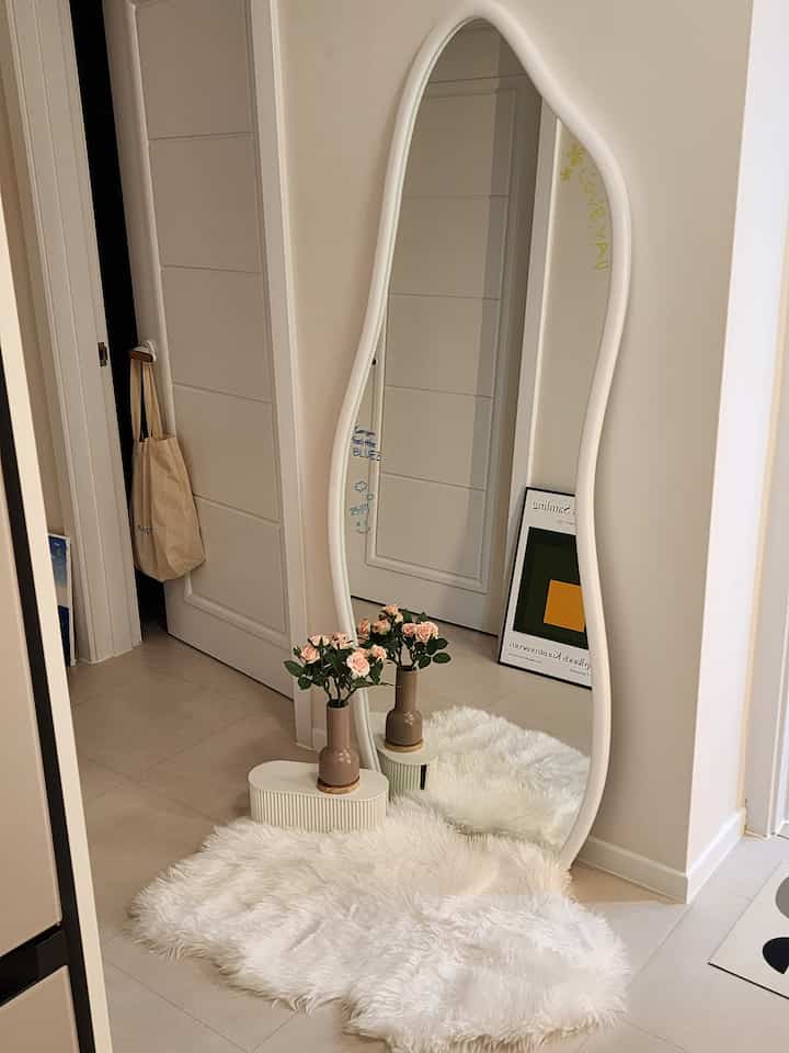 白を基調とした玄関に大きな曲線型の全身鏡とベージュの花瓶、白いラグが配置されたモダンで落ち着いたインテリア