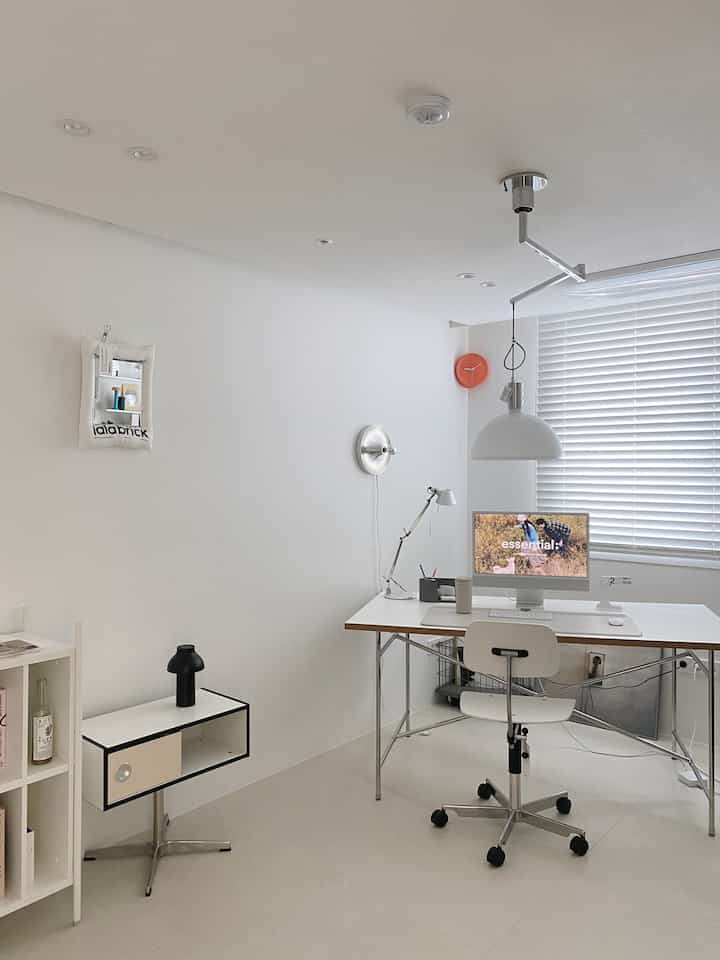 A minimal white-toned study featuring a clean desk, chair, and blinds, creating a tidy workspace