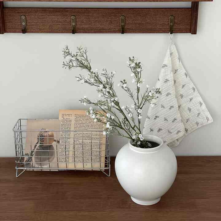 Wood tone table featuring a white vase with artificial flowers and a patterned white towel hung on the wall, creating a cozy decor space