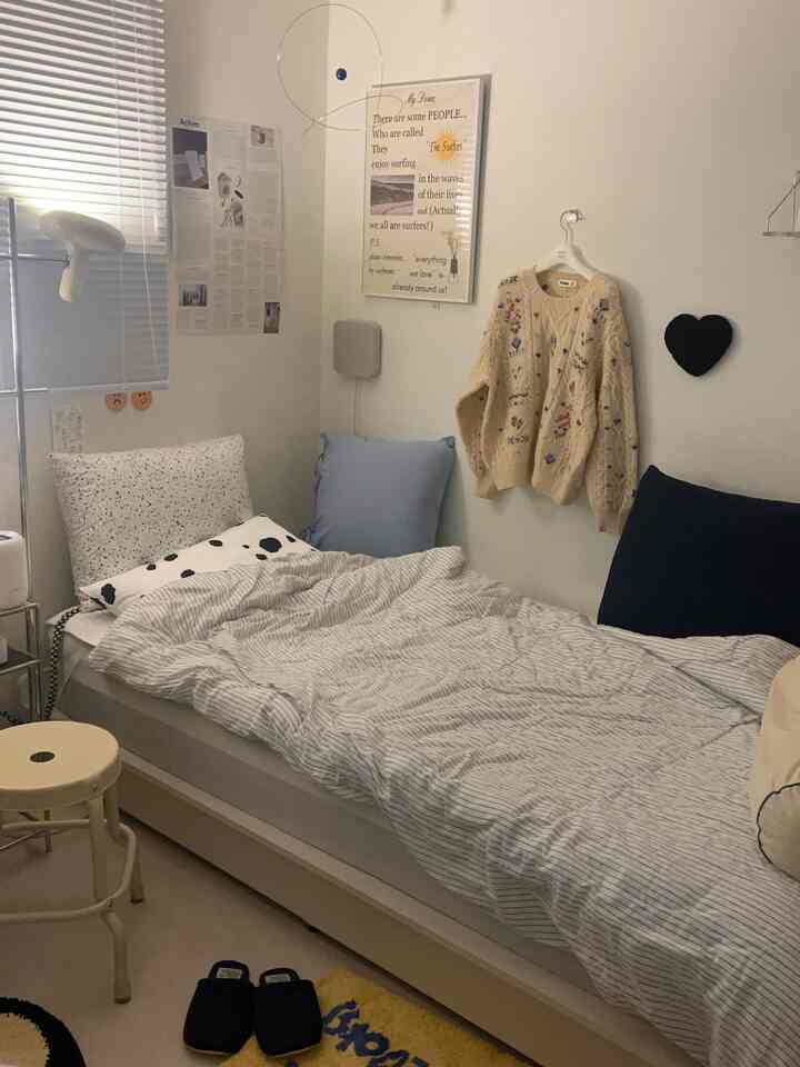 ホワイトとブルーを基調とした一人暮らしの寝室、コンパクトで居心地の良い空間にベッドとスツール、ヴィンテージ風セーターの壁飾りが配置された雰囲気のあるインテリア