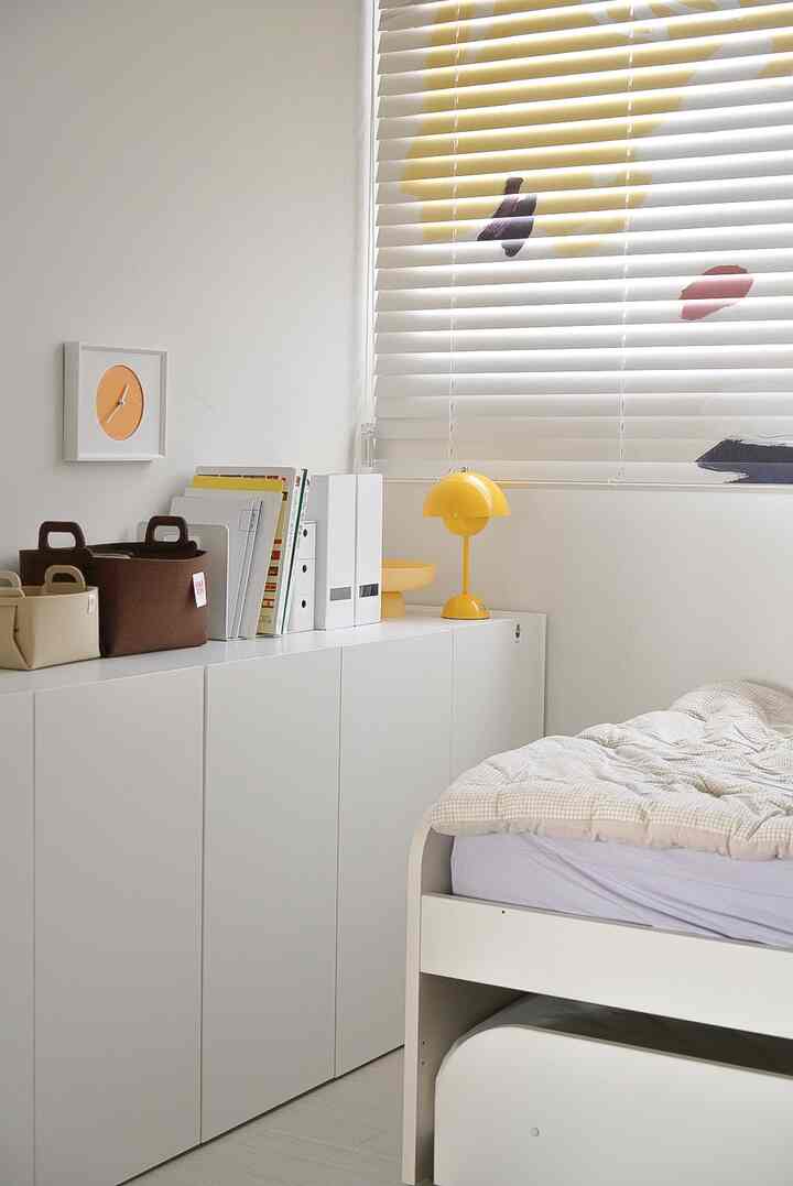 白を基調とした子供部屋、整然とした収納キャビネットと黄色いテーブルランプが配置された印象的な空間