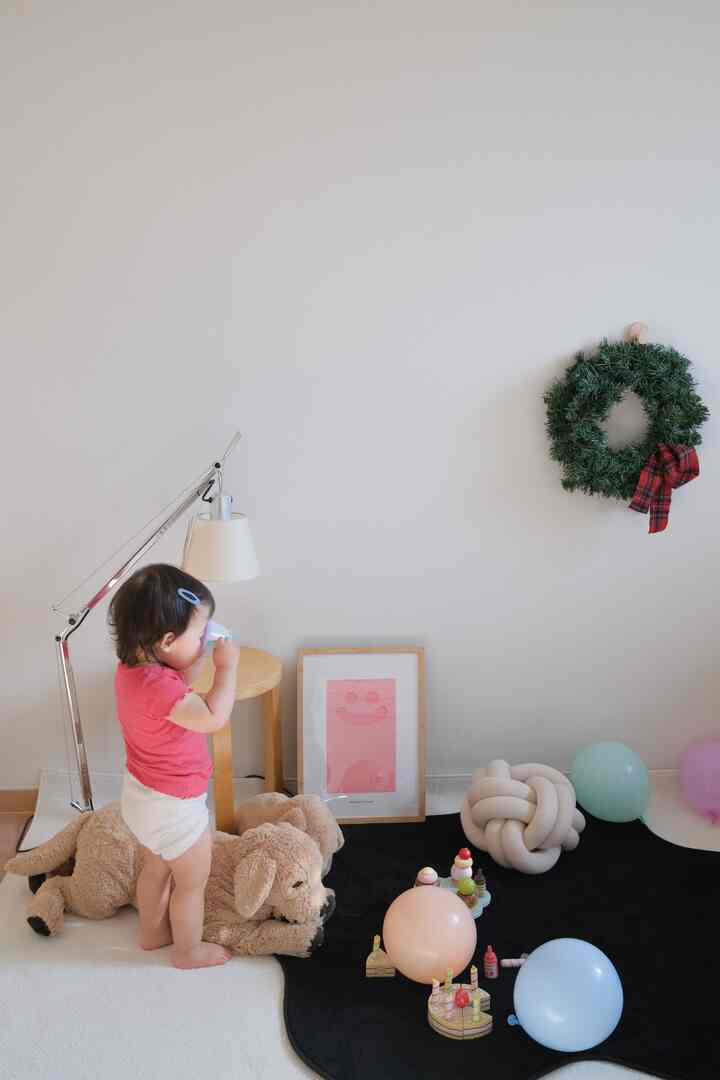 白い壁と黒いラグが調和した子供部屋で、赤ちゃん、ぬいぐるみの犬、テーブルランプ、額縁が配置された印象的な空間
