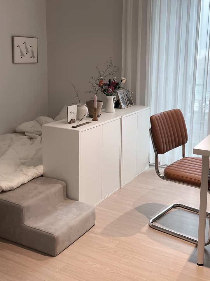 Bright white-toned studio apartment featuring desk, brown chair, and centrally placed white storage divider creating a cozy interior