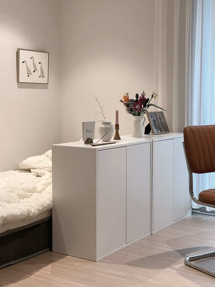 Bright white-toned bedroom featuring a white storage cabinet center, bed left side, and brown chair right, with a clean modern aesthetic