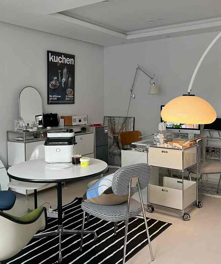 明るいグレーの壁のモダンなダイニングルーム、中央の丸テーブルにメディアエアフライヤーと飲み物が置かれ、多様な椅子と照明が配置された印象的な空間