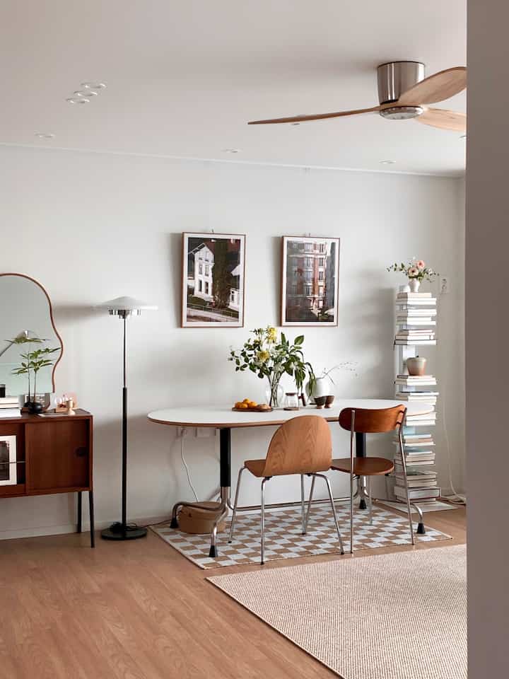 Minimal living room in white and wood tones featuring a white dining table, wooden chairs, rugs, picture frames, and floor lamp in harmonious arrangement