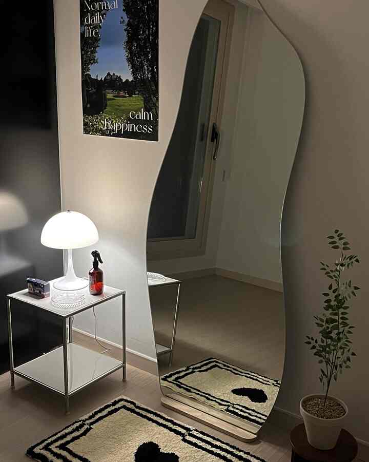 白と茶色のトーンの狭い部屋、曲線型の全身鏡とサイドテーブル、ムードランプが配置されたモダンで居心地の良い空間