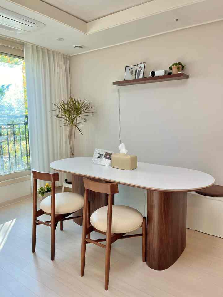 White and natural wood tone living room featuring white dining table, wooden chairs, large window, and plant in a simple cafe-style atmosphere