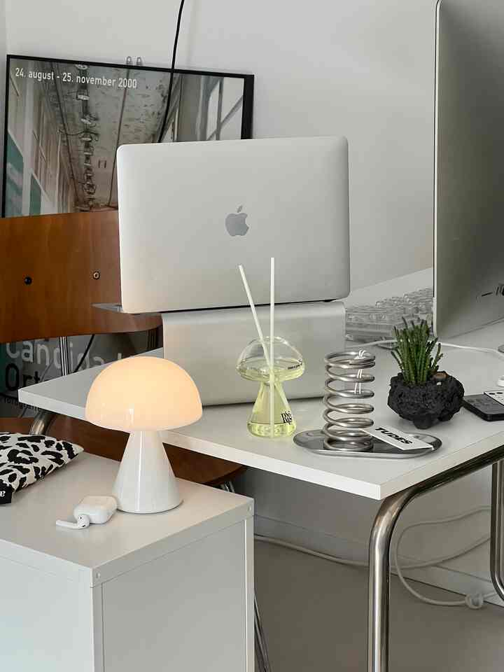白とシルバーのトーンのホームオフィス、モダンなデスクに配置されたMacBook、ランプ、ディフューザーのある印象的な空間