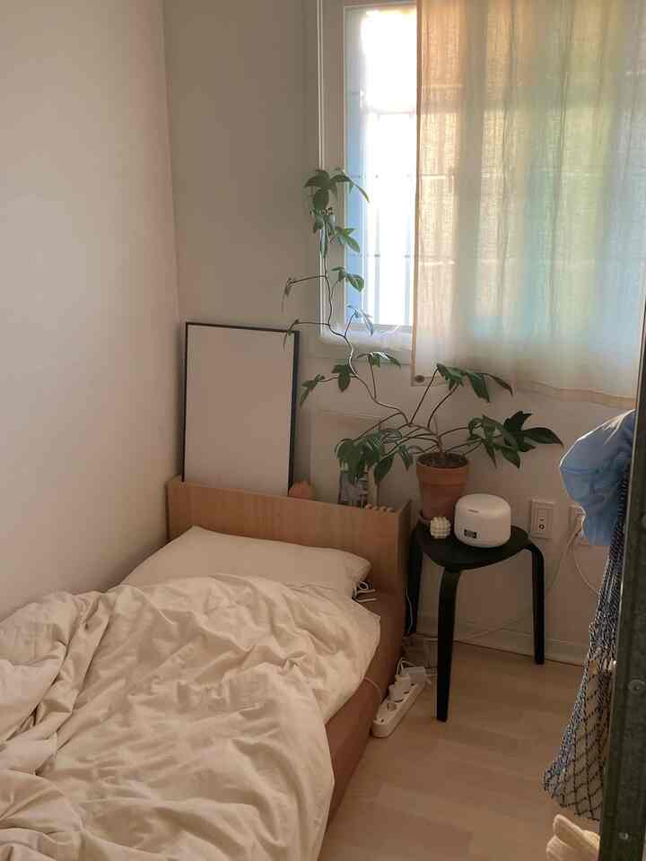 ナチュラルな色調の寝室、木製ベッドとベージュの寝具、小さな黒いナイトテーブル、窓際の観葉植物が配置された雰囲気のあるインテリア