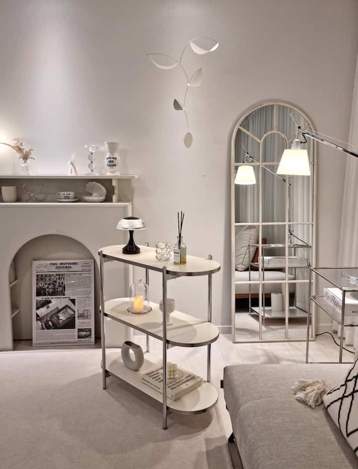 White-toned living room featuring shelves, large mirror, modern lighting, and interior accessories creating cozy and clean atmosphere