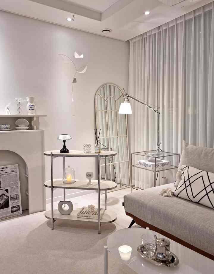 White-toned living room featuring a 3-tier shelf, full-length mirror, modern lighting, and sofa in a clean minimalist interior