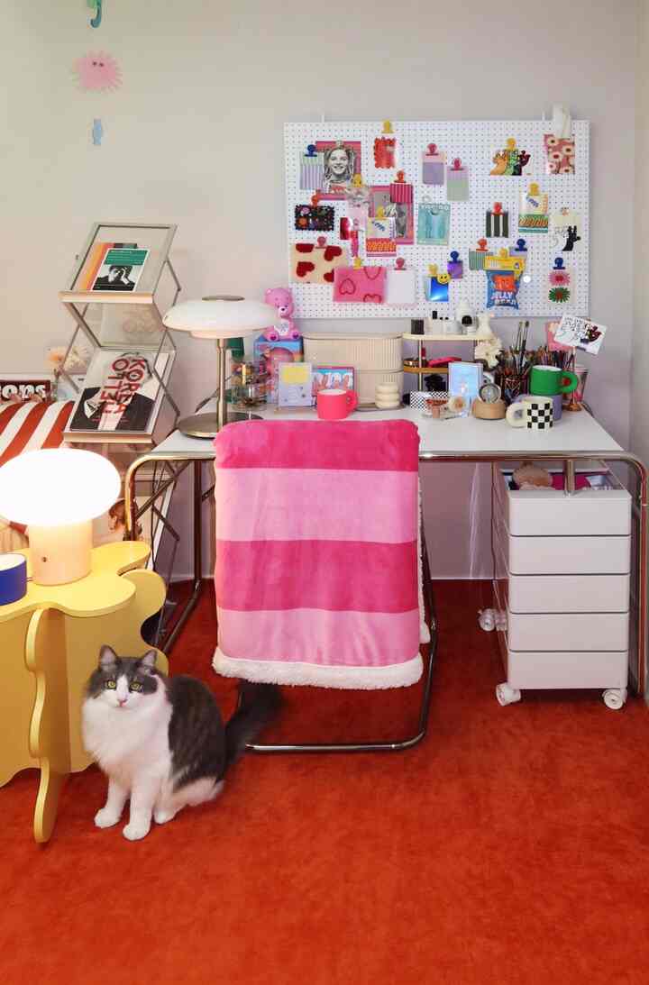 白い壁とピンクのブランケットがあるミッドセンチュリーモダン風のデスクスペース、鮮やかな赤いカーペットの上に猫が座っている雰囲気のあるインテリア