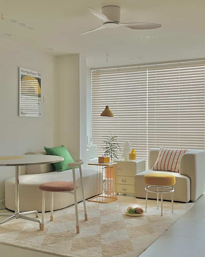 Bright white-toned living room featuring white sofas, beige rug, yellow stools, and green cushions arranged in a clean, natural space