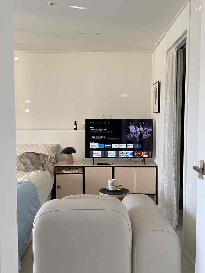 ホワイトとベージュの色調の寝室、左側のベッドに猫がいて、シンプルな印象のテレビ台付きキャビネット配置の空間