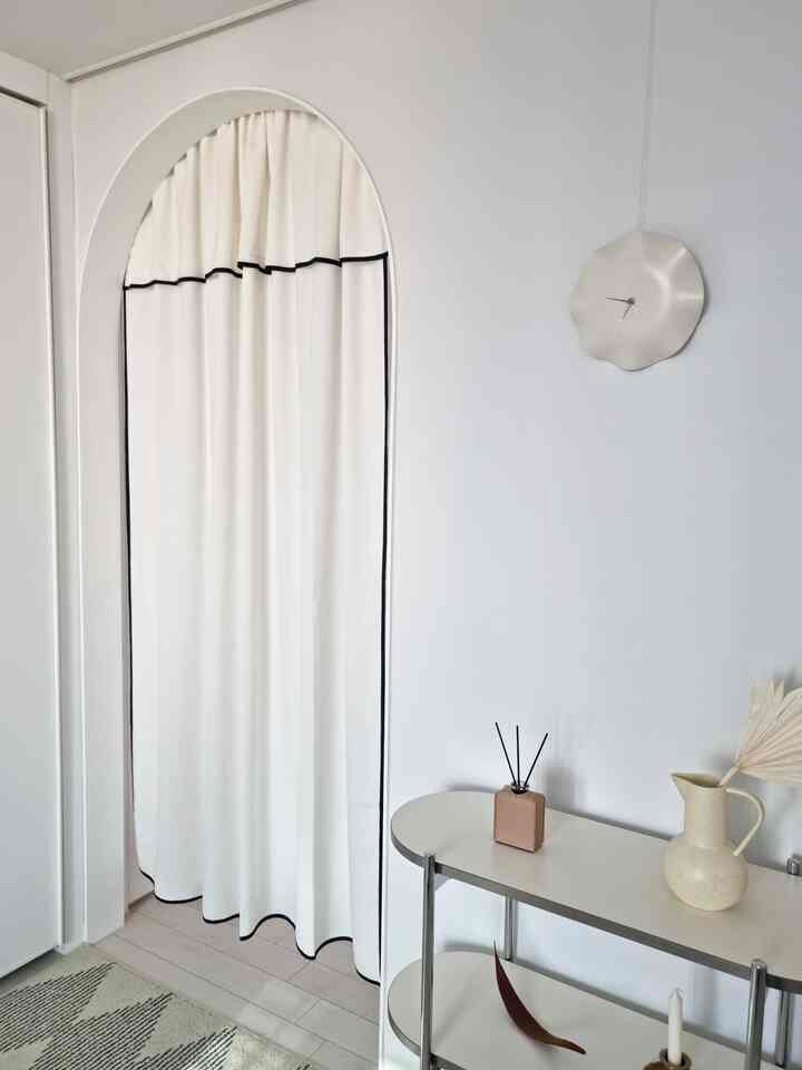 白を基調とした寝室空間、アーチ型の入り口に白いカーテンが掛けられ、棚とユニークな壁時計が配置された雰囲気のあるインテリア