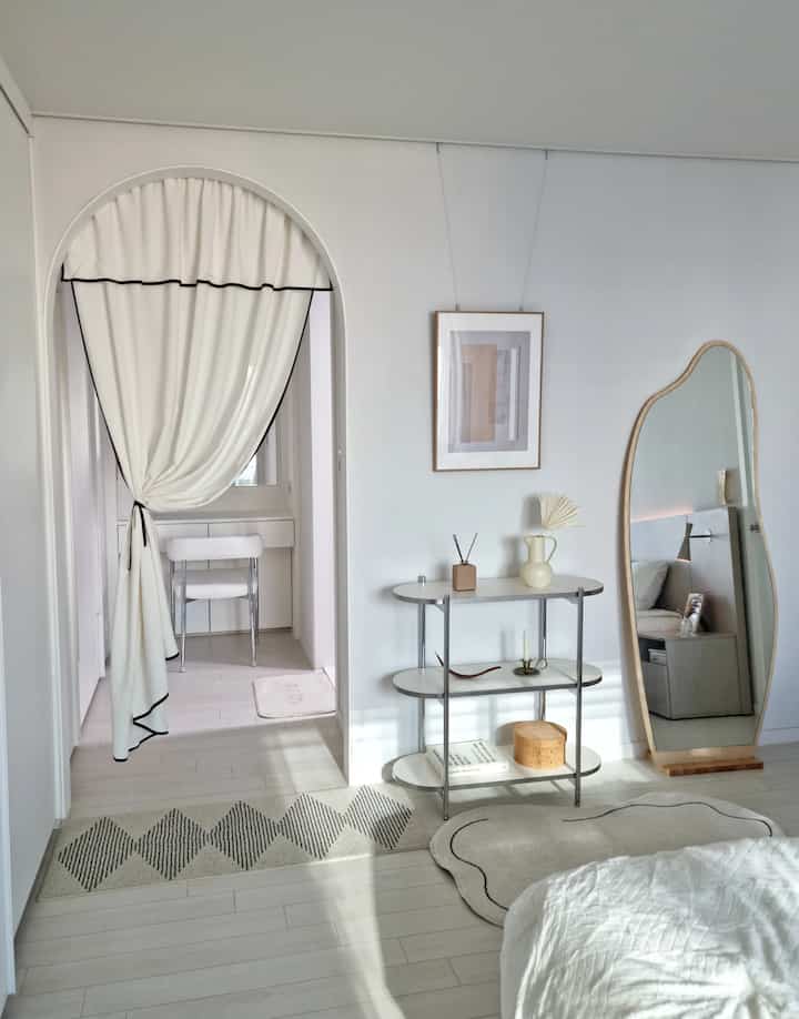 White-toned bedroom featuring an arched partition with French-style curtain divider and modular shelf, creating a cozy interior