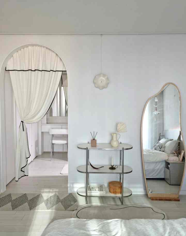 White-toned bedroom featuring a soft curtain divider, asymmetrical full-length mirror, three-tier shelf, and wall clock in a clean mid-century modern style