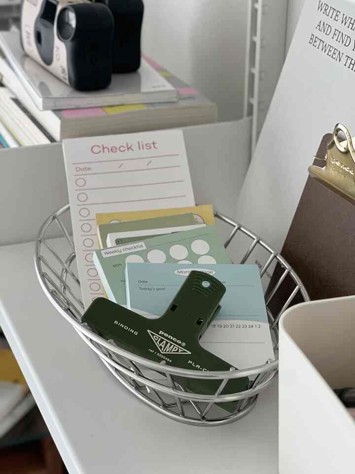 Silver metal basket on a white desk holding green clip and assorted sticky notes, organized and clean workspace