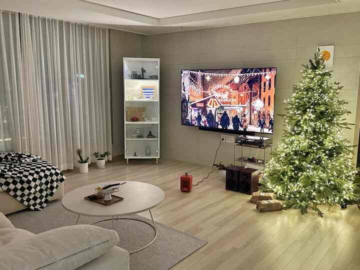 ホワイトとナチュラルトーンのリビングルーム、クリスマスツリーとガラス陳列棚、壁掛けテレビが配置されたモダンな空間