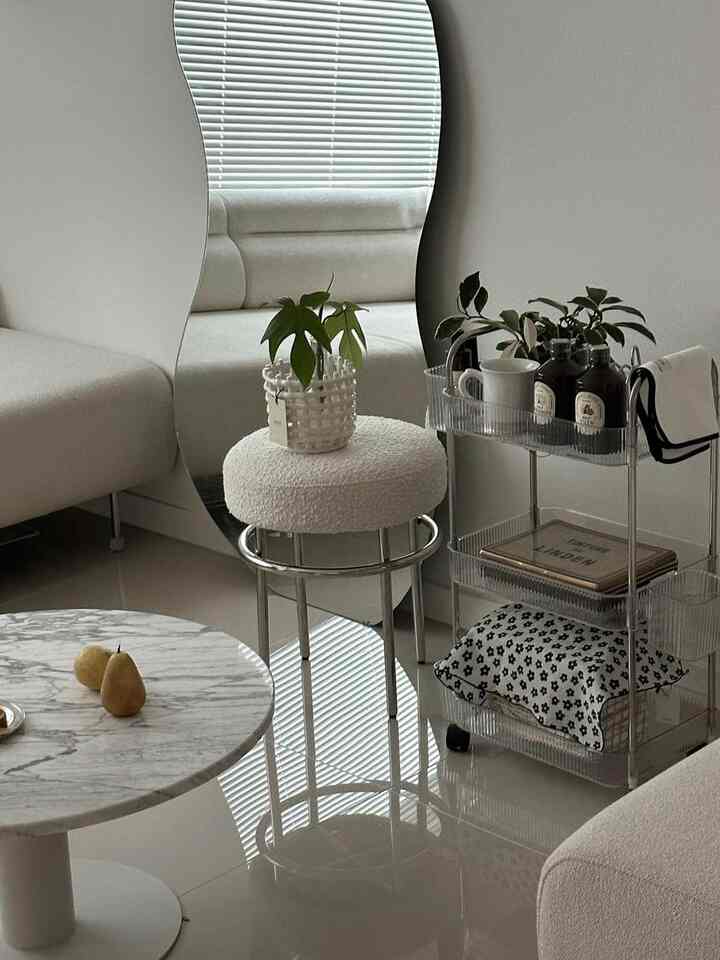 明るいホワイトトーンのリビング、透明なアクリル製の3段トロリーと曲線的な鏡が配置されたシンプルでモダンな印象のインテリア