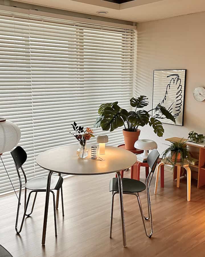 白いブラインドを背景にしたリビング、丸いテーブルとアルノチェア、多様な植物が配された温かみのあるナチュラルなインテリア