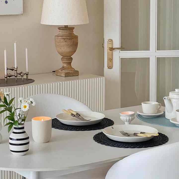 White and beige toned dining room featuring a white dining table, plates, black placemats, and lit candles creating a warm ambiance