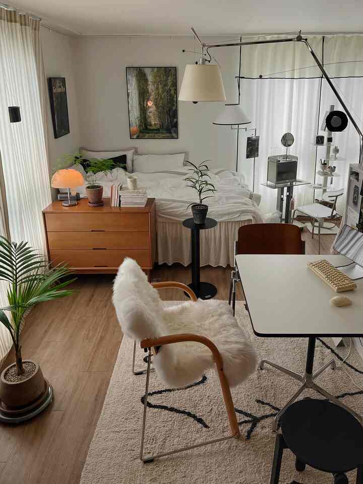 ホワイトと木目調が調和したワンルームの寝室兼ホームオフィス、植物と多様な照明が温かみのある雰囲気を演出