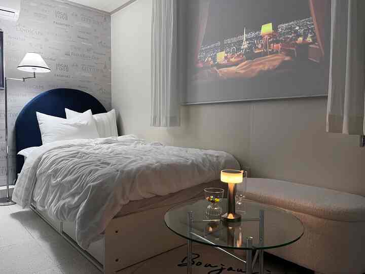 白とネイビートーンが調和する6坪のワンルーム寝室、ベッドとガラステーブル、壁に映るプロジェクター画面が配置された落ち着いた空間