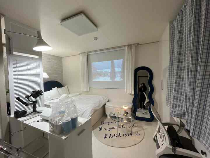 White and beige toned studio apartment featuring a bed, curvy blue mirror, desk, and projector screen in a clean, minimal setting